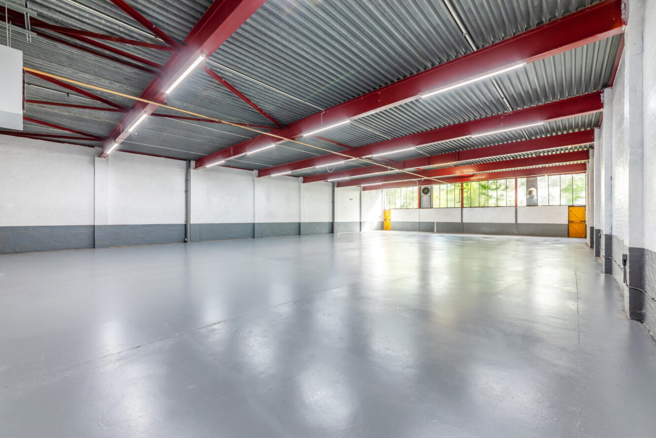 Large empty industrial warehouse with gray concrete floor, white and gray walls, red steel beams, and fluorescent ceiling lights. Windows and yellow doors are visible at the far end.