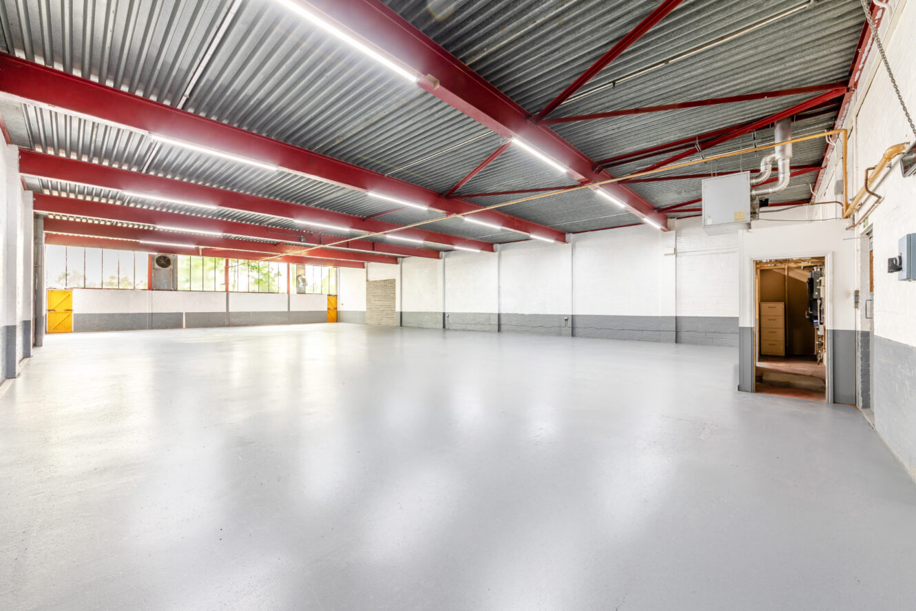 Spacious, empty industrial warehouse with gray floors, white and gray walls, exposed red ceiling beams, fluorescent lighting, large windows, and an open doorway to a small adjoining room.