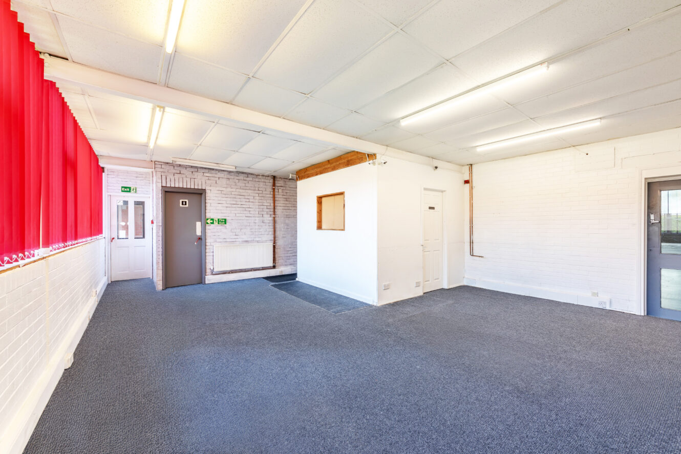 Empty room with white brick walls, grey carpet, red vertical blinds on the left, fluorescent ceiling lights, and a small window in an interior partition wall.