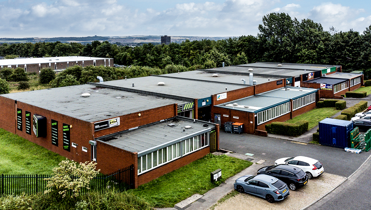 A row of single-story brick commercial buildings with flat roofs, surrounded by parked cars, trees, and distant fields under a partly cloudy sky.