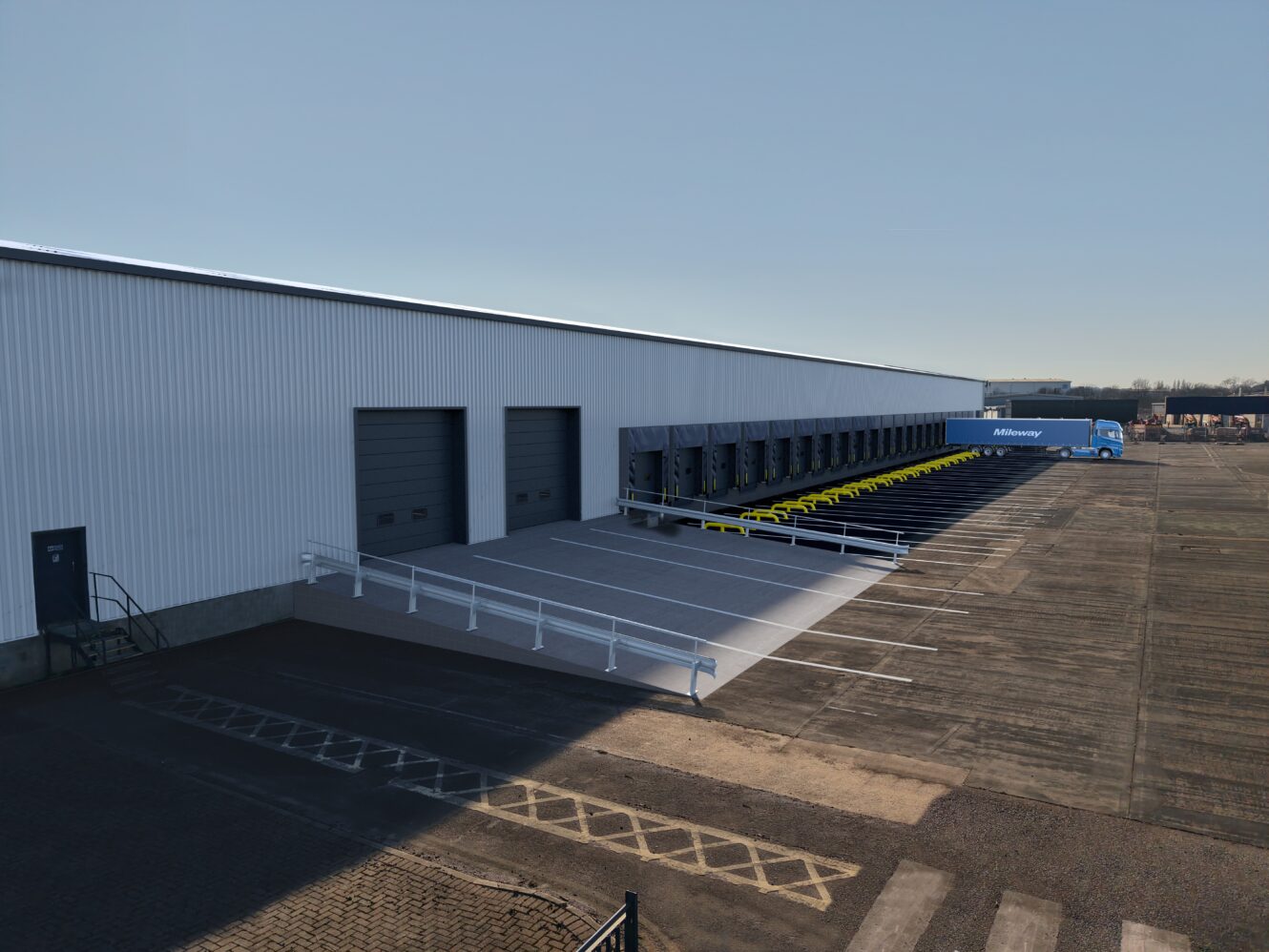 A large warehouse loading dock with multiple loading bays, some with docked trailers, and a blue delivery truck parked on the far right.