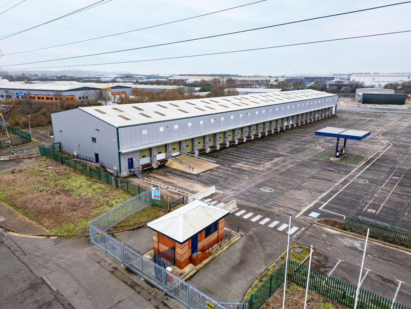A large industrial warehouse with multiple loading bays, a security gatehouse, and an empty parking lot surrounded by fencing.
