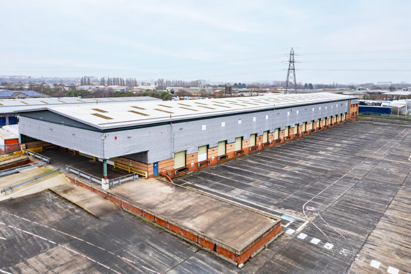 A large industrial warehouse with multiple loading bays and an adjacent empty parking lot, situated in an industrial area with power lines in the background.