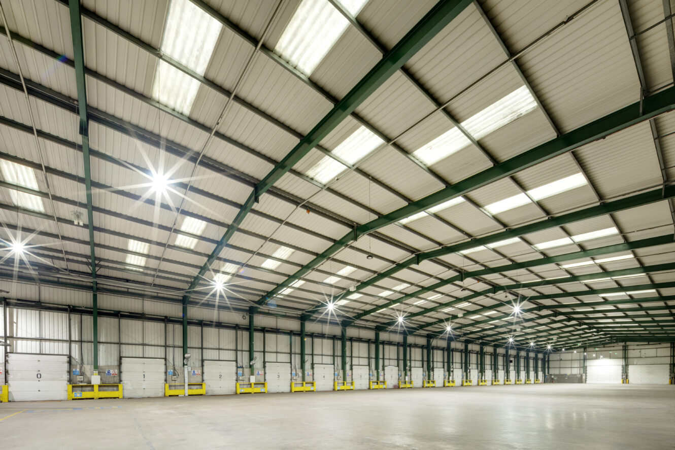 Spacious, empty warehouse interior with a high ceiling, metal beams, bright overhead lights, and multiple loading dock doors along one wall.