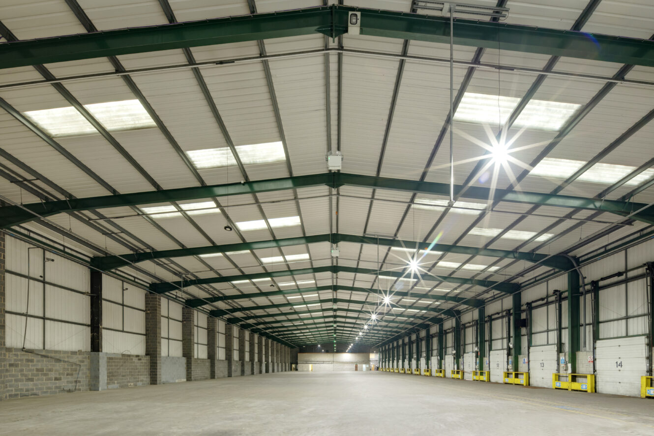 Interior of a large, empty warehouse with high ceilings, skylights, green steel beams, concrete floors, and numbered yellow loading bays along one wall.