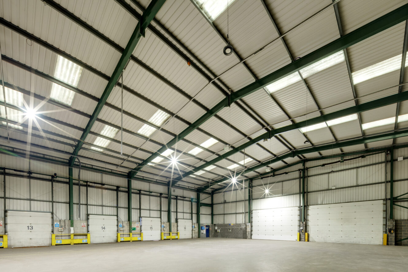 Interior view of a large, empty warehouse with metal beams, overhead lights, and several loading dock doors along one wall.
