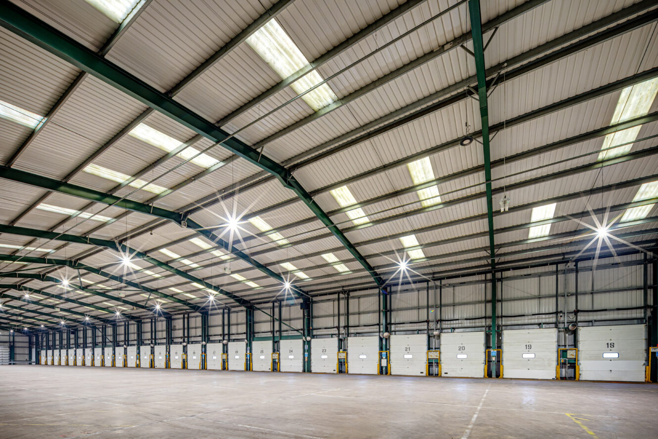 Large, empty warehouse interior with high ceilings, metal beams, skylights, and a row of numbered loading bay doors along one wall.