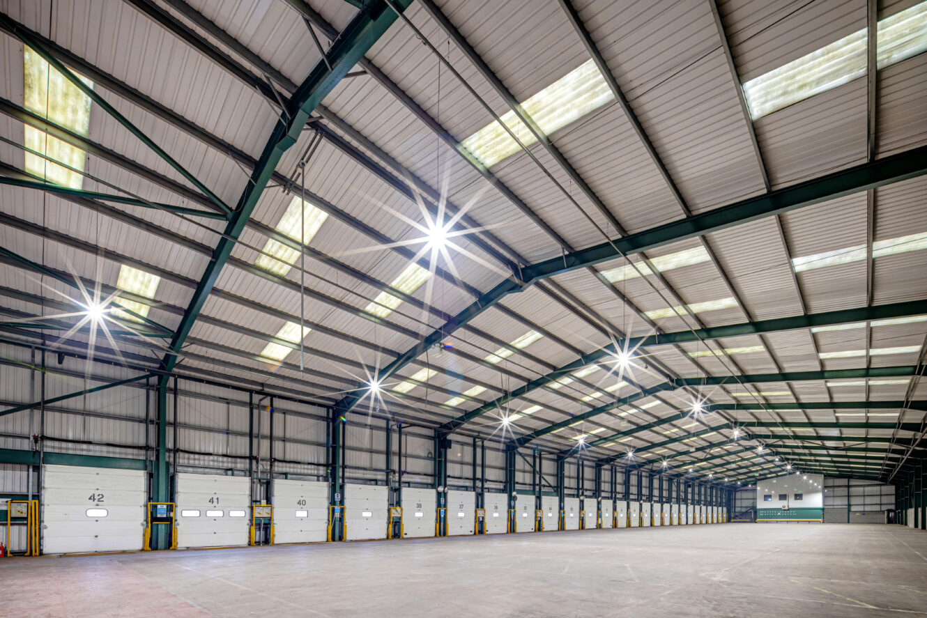 Interior of a large, empty warehouse with high ceilings, overhead lights, and numbered loading bay doors along one wall.