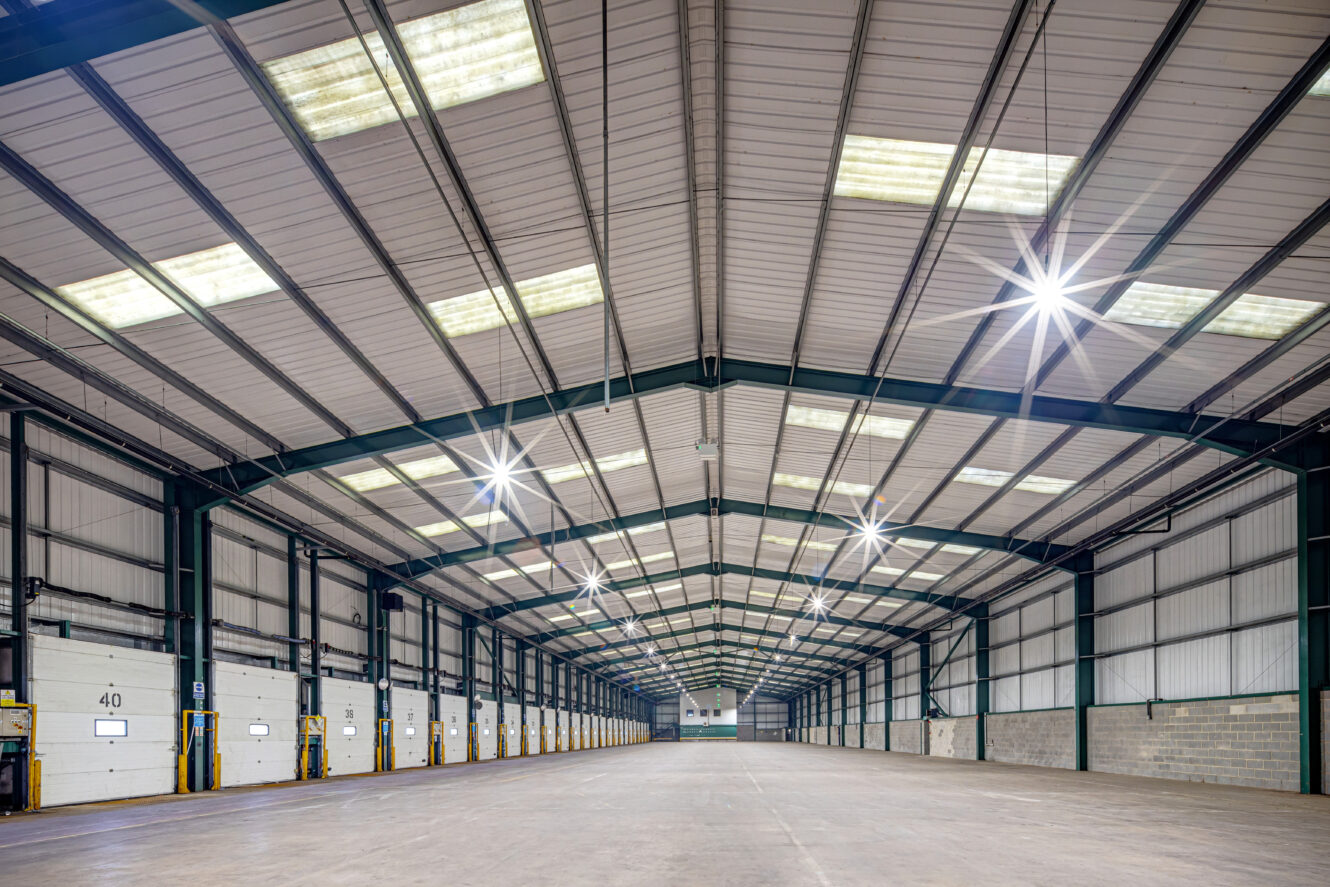 Large, empty warehouse with high ceilings, overhead lighting, and multiple loading dock doors along the left wall.