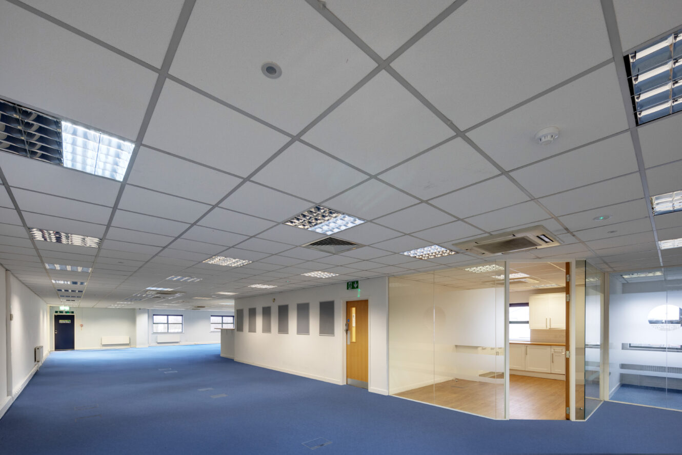 Empty office space with blue carpet, white ceiling tiles, overhead fluorescent lighting, glass-walled meeting room, and kitchen area visible in the back.