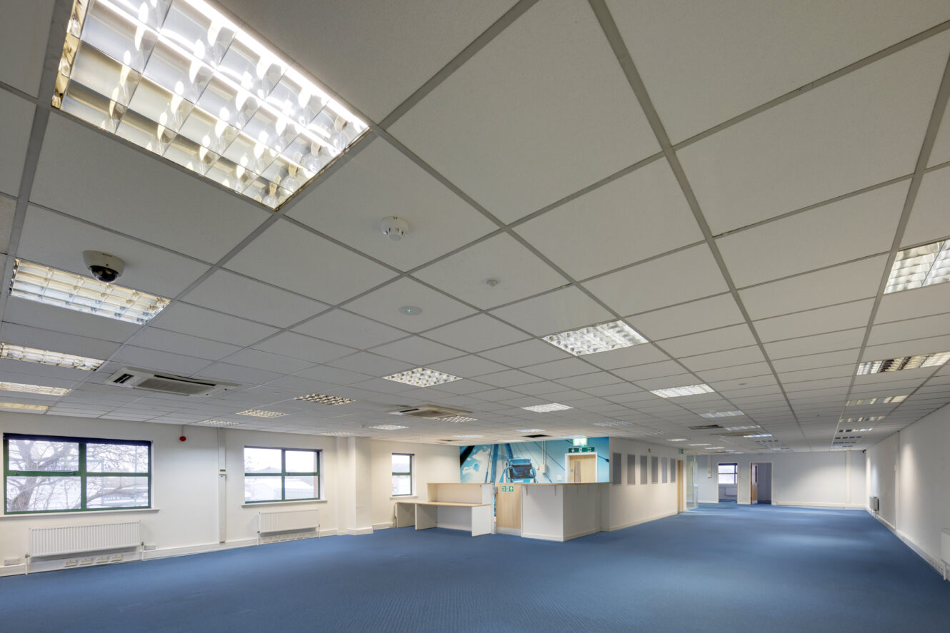 Large empty office space with blue carpet, white ceiling tiles, fluorescent lights, several windows, and a few partitioned areas near the back.