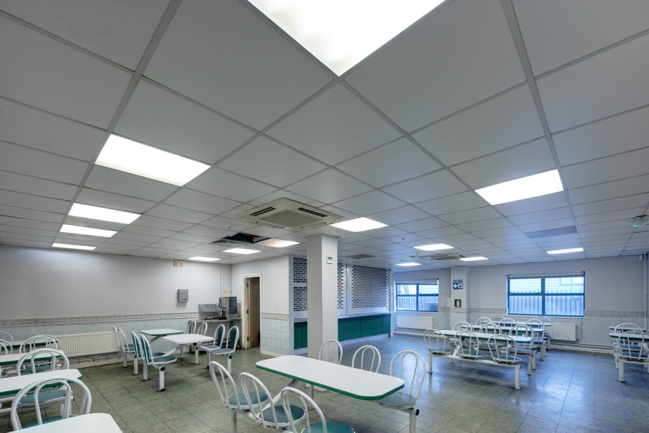 An empty cafeteria with white tables, metal chairs, tiled floor, and fluorescent ceiling lights. Windows line the back wall, and a serving area is visible on the right.