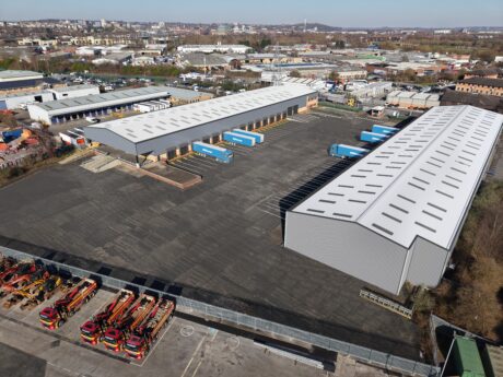 Aerial view of a large industrial warehouse complex with loading docks, several blue trucks, and equipment parked in an open lot. Urban buildings and greenery are in the background.