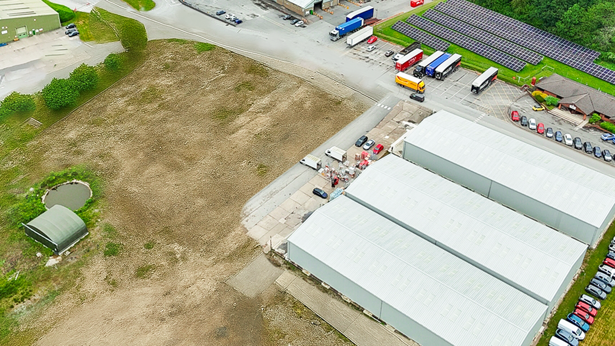 Aerial view of an industrial site with warehouses, parked trucks, cars, solar panels, a pond, and adjacent open land.