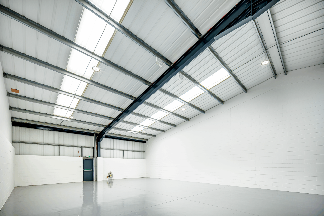 Empty industrial warehouse with a clean, polished floor, white brick walls, metal beams, large windows in the ceiling, and a single exit door.