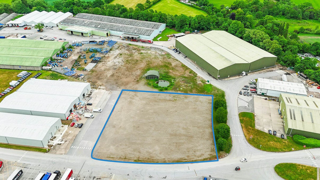 Aerial view of an industrial area with a large, vacant, rectangular plot of land outlined in blue, surrounded by several warehouses and greenery.