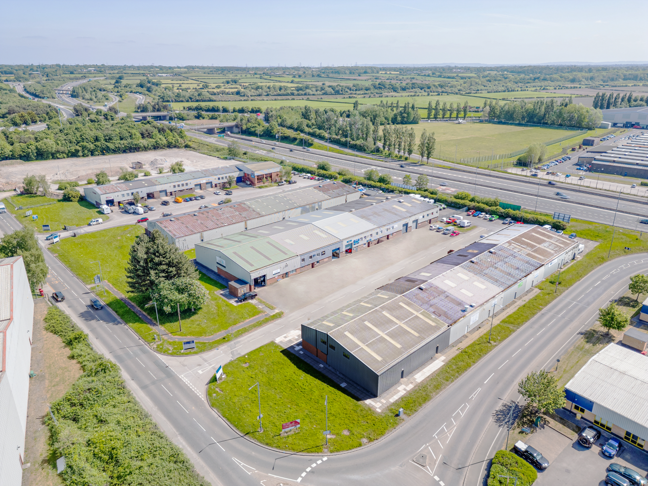 Aerial view of an industrial estate with multiple warehouses, parking areas, and surrounding roads, set near a highway and open green fields.