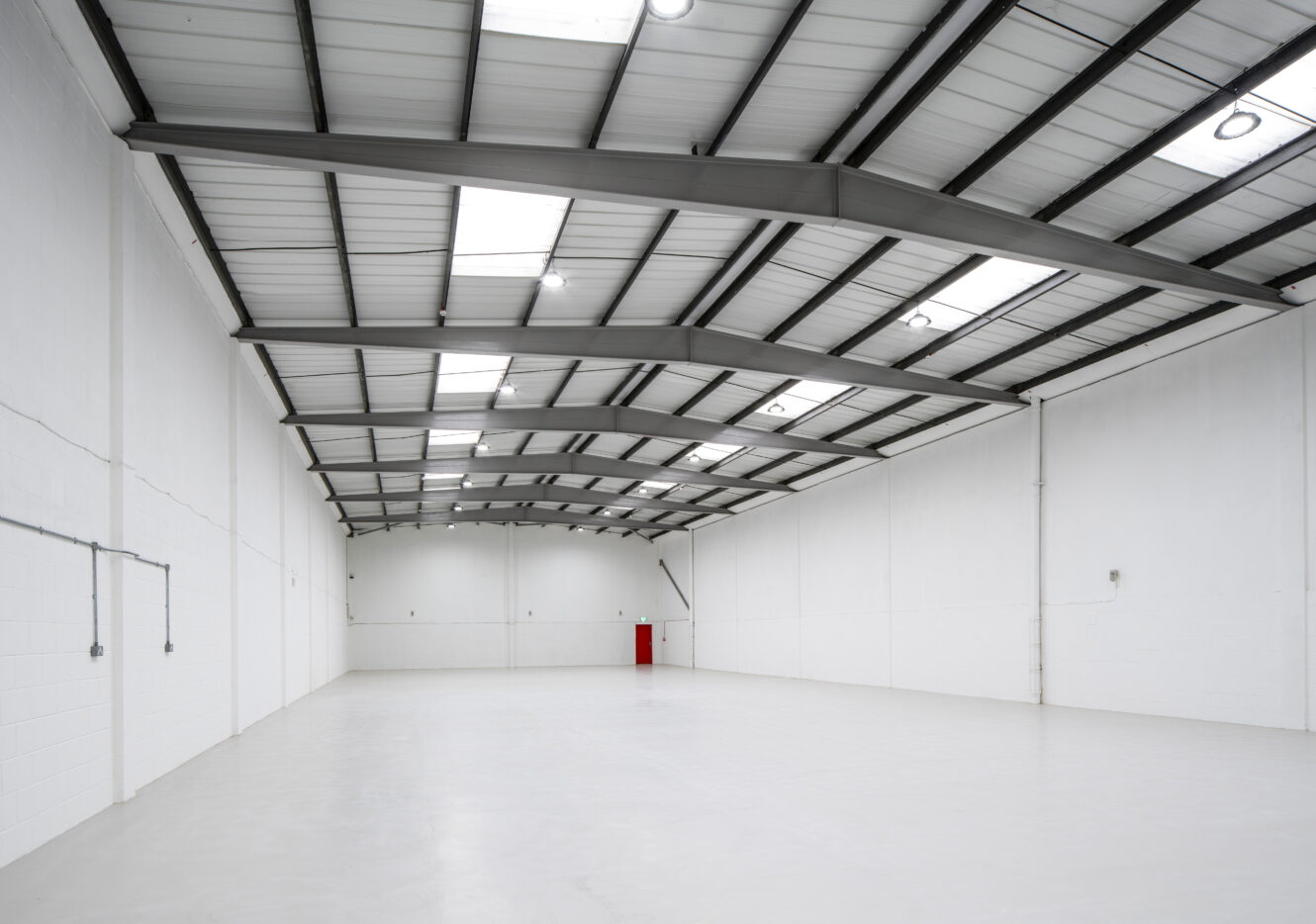 Large, empty industrial warehouse with white walls, concrete floor, high ceiling, and exposed beams; overhead lights are on and a red fire hose station is visible at the far end.