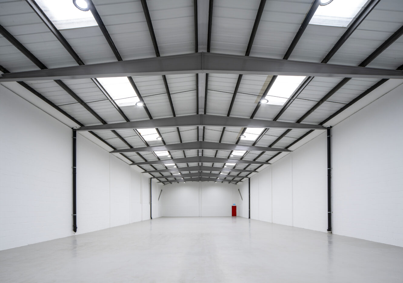 Empty industrial warehouse with a high ceiling, white walls, polished concrete floor, skylights, and a single red fire extinguisher at the far end.