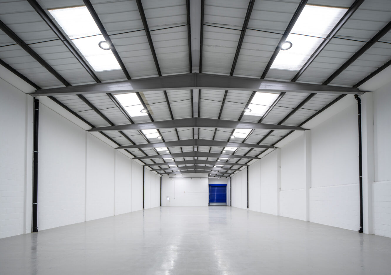 Empty industrial warehouse with white walls, high ceilings, metal roof beams, and skylights; a large blue door is visible at the far end.