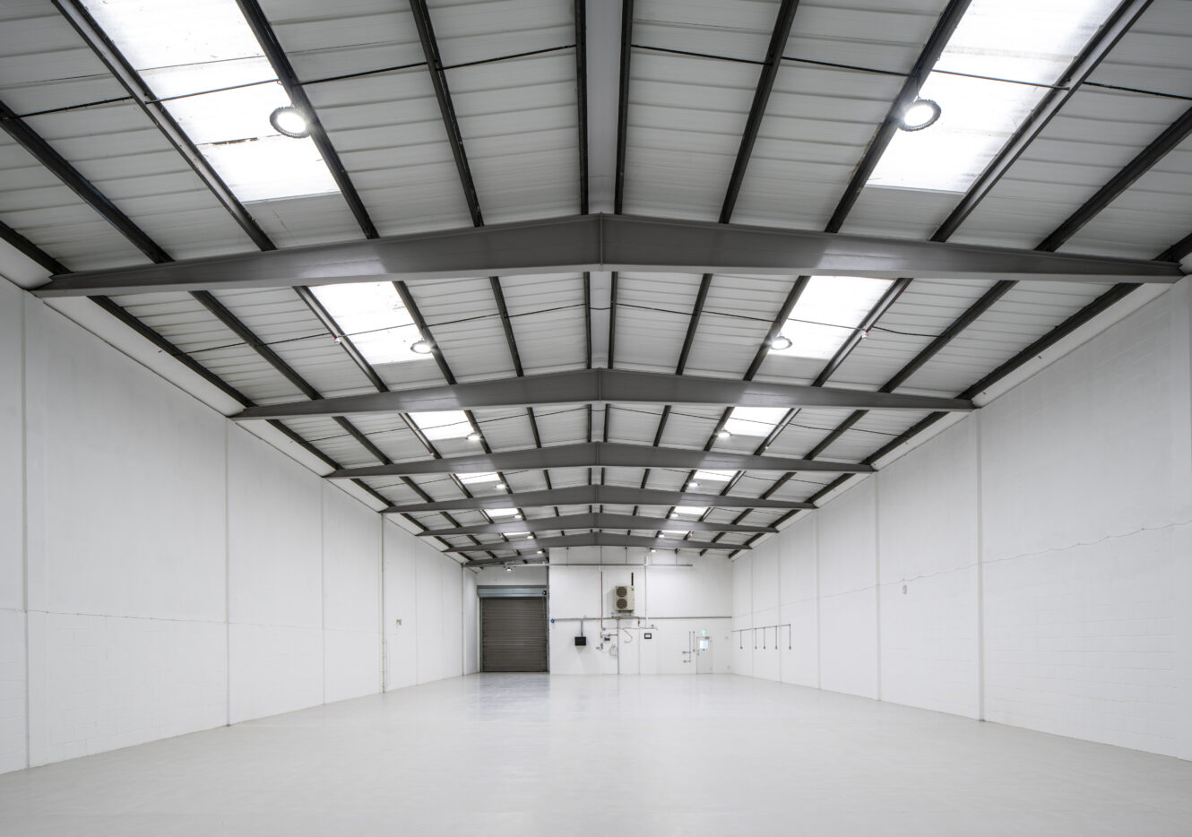 Interior of an empty warehouse with high ceilings, white walls, overhead lights, and a large shutter door at the far end.