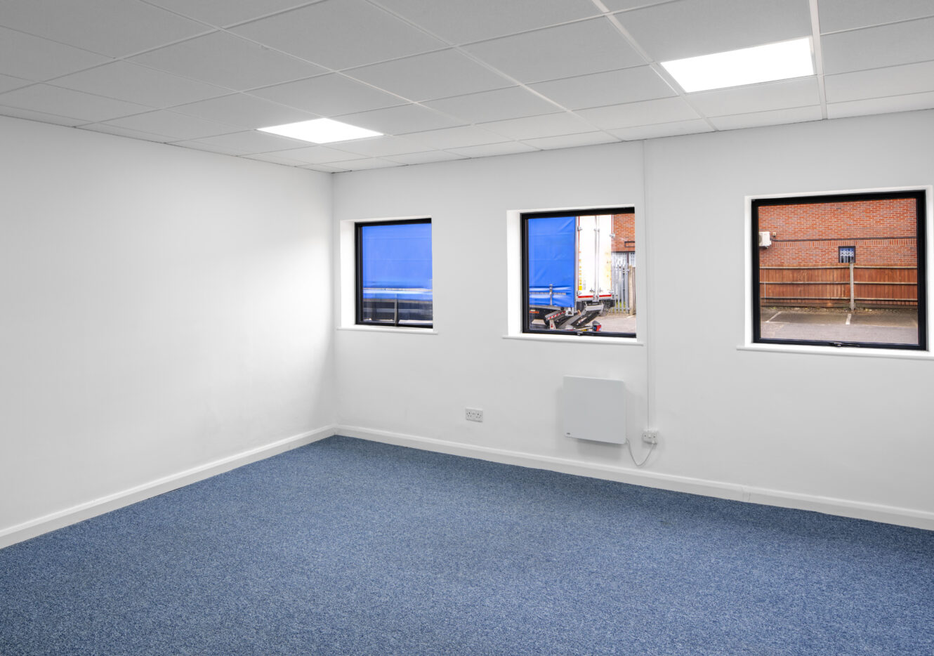 Empty office room with white walls, blue carpet, three square windows, and ceiling lights. A parking lot and brick wall are visible through the windows.