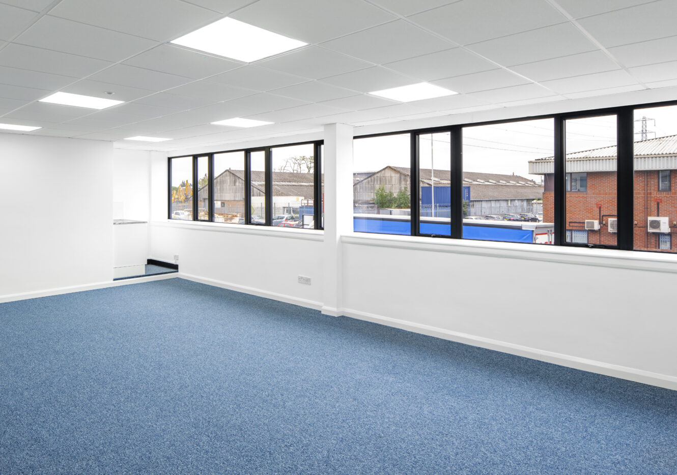 Empty office space with blue carpet, white walls, ceiling lights, and large windows overlooking an industrial area.