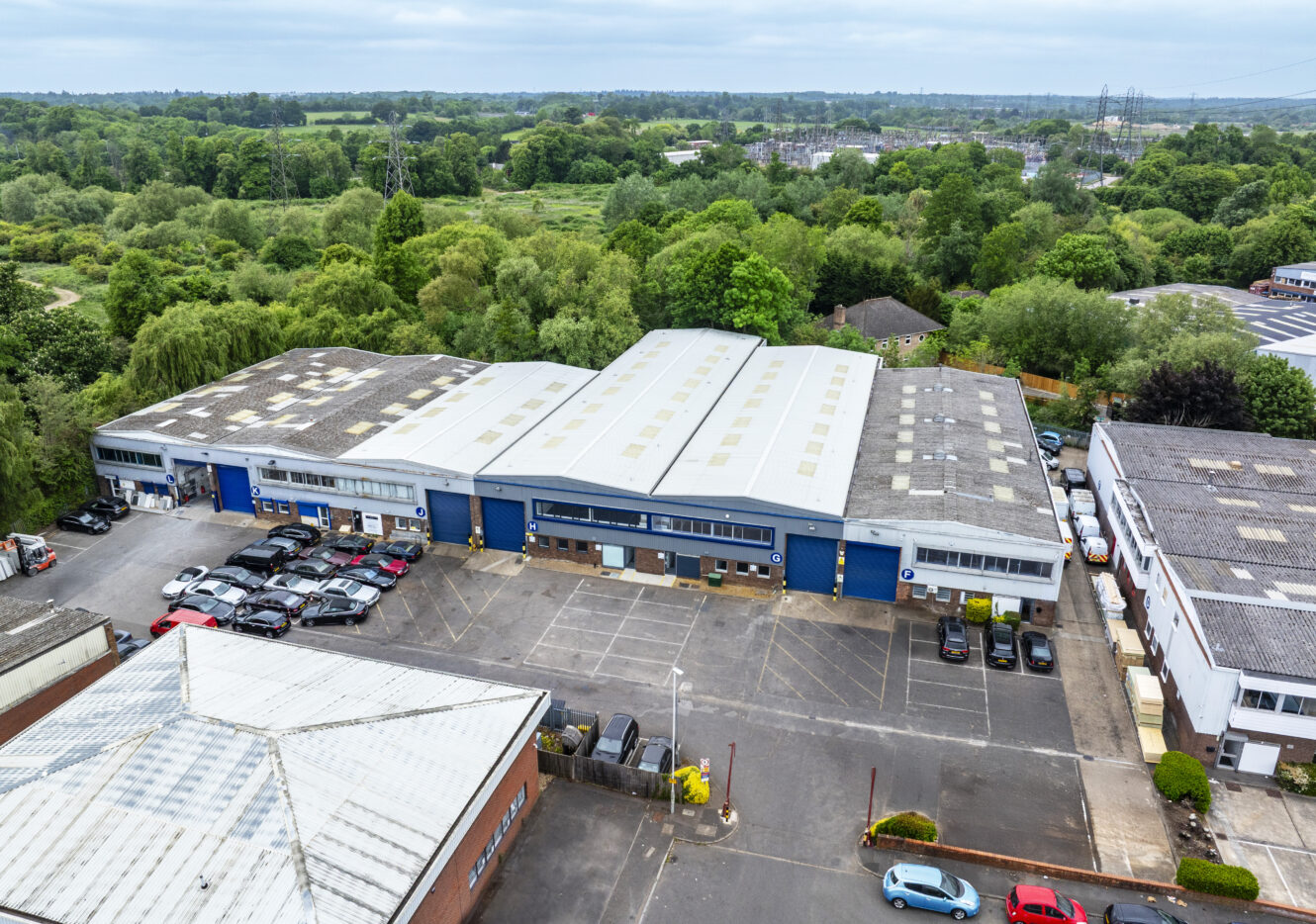Aerial view of a commercial industrial building with a large parking lot, surrounded by trees and other similar structures. Several cars are parked near the entrance.