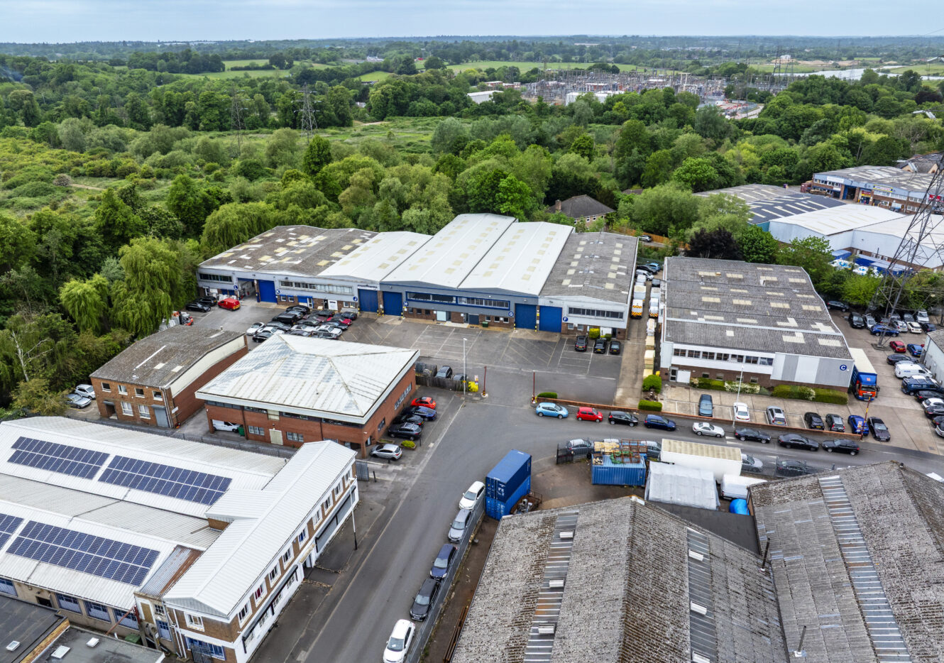 Aerial view of an industrial estate with multiple warehouses, office buildings, vehicles parked outside, and green trees and fields in the background.