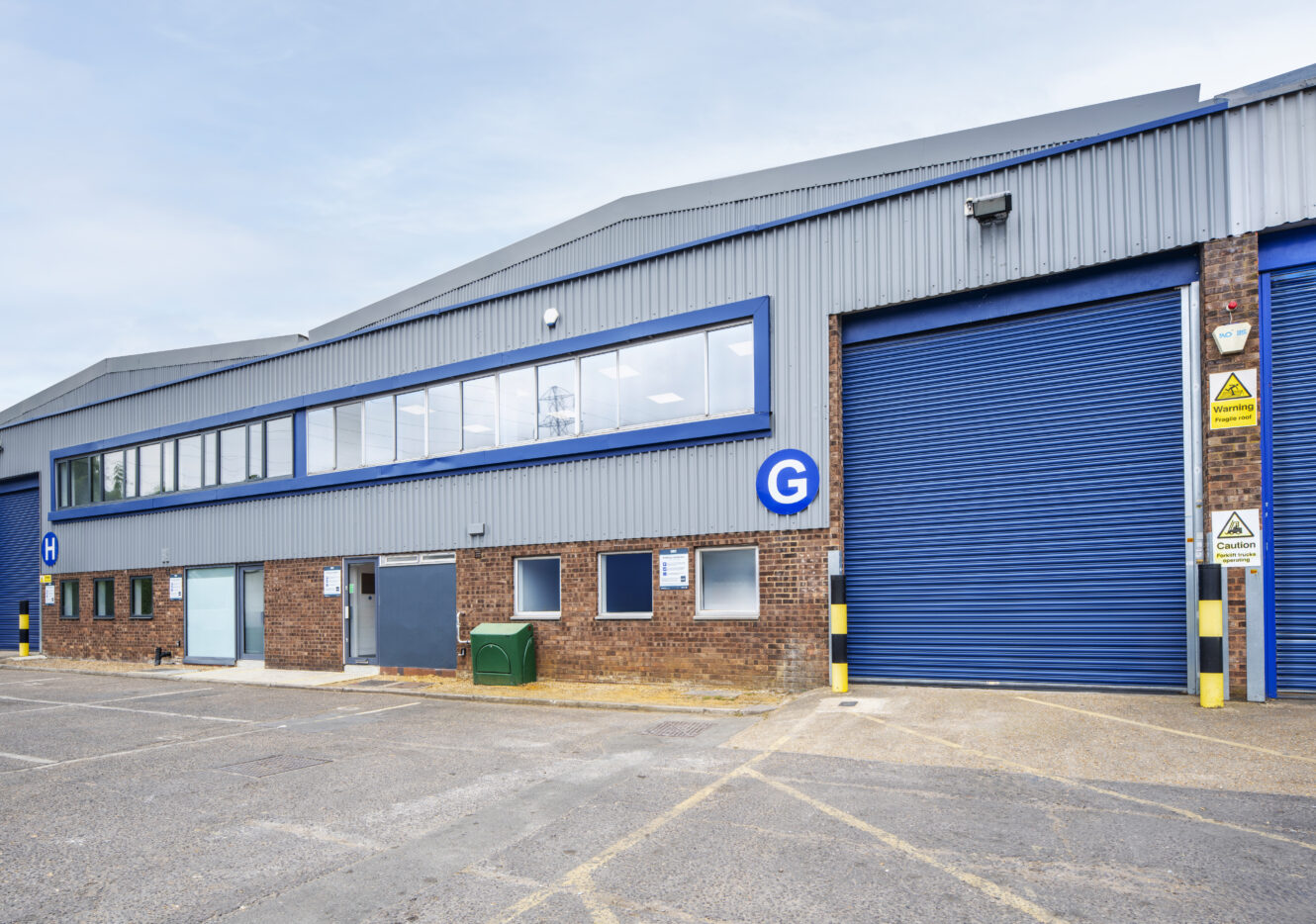 Exterior of an industrial warehouse with blue doors, brick and metal siding, and a large G sign. The area in front is empty and paved.