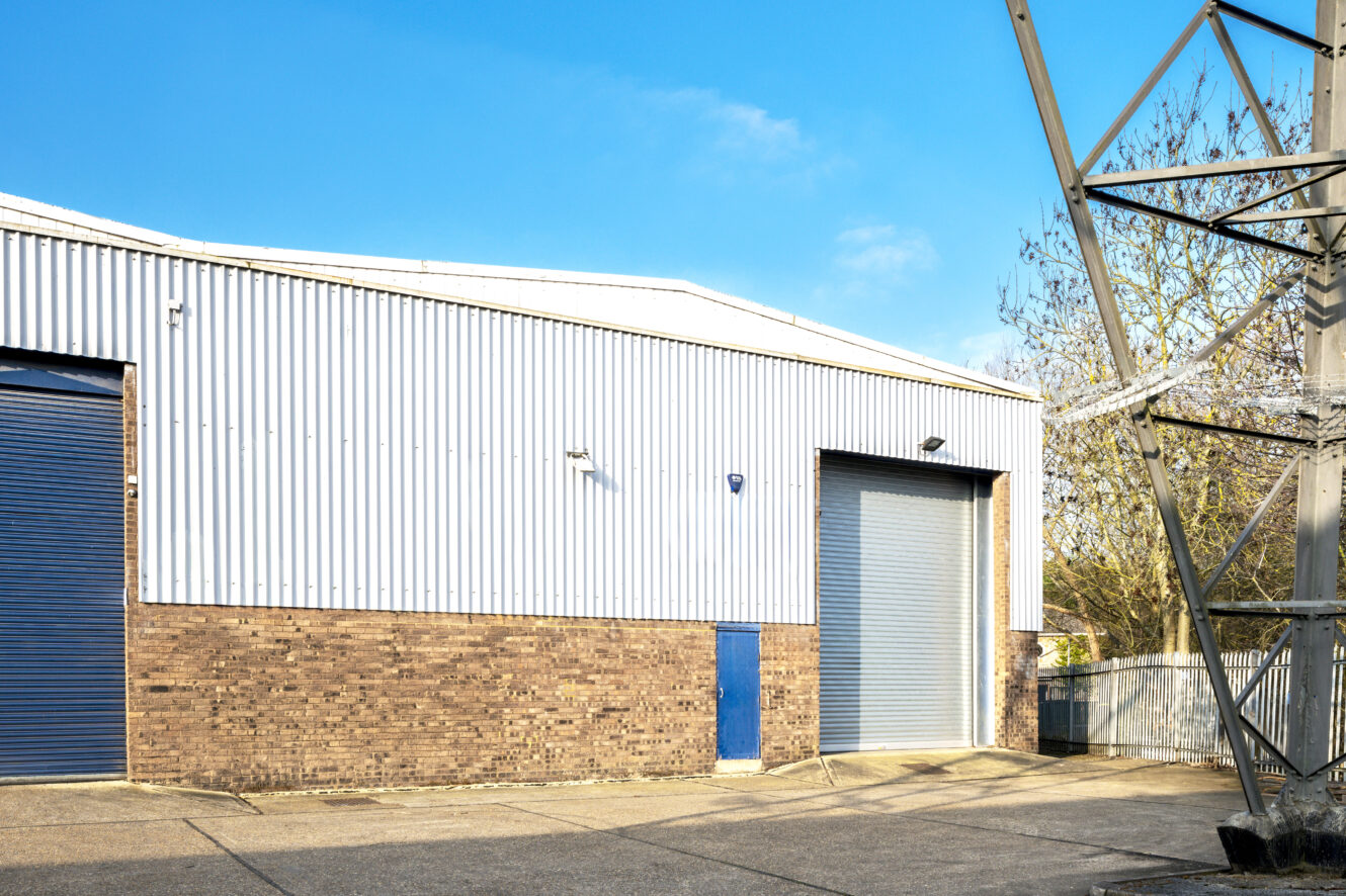 A large industrial warehouse with brick and metal siding, blue roller doors, and a security camera, under a clear blue sky.
