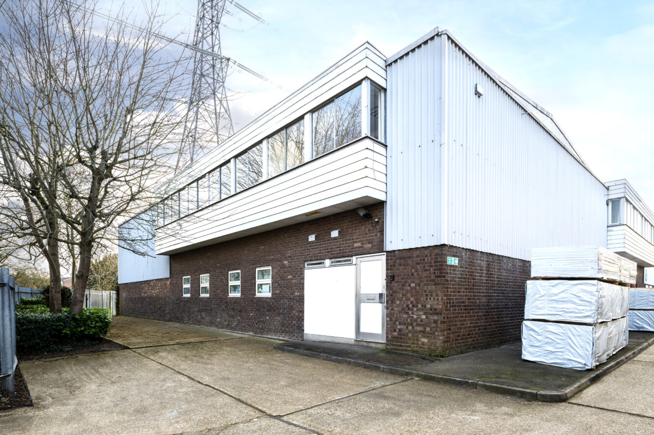 A two-story industrial building with brick lower walls and metal upper siding, surrounded by paved ground and stacks of wrapped materials.