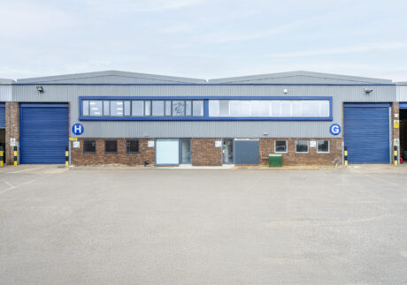 Industrial warehouse building with two large blue roller doors, brick facade, upper row of windows, and letters H and G marking each door.