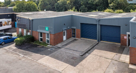 A grey industrial warehouse with two large blue roller doors, a white entrance door, and a green 5 sign above the main entrance, surrounded by trees and parked cars.