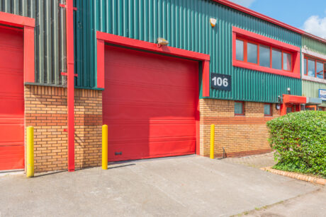 A red industrial garage door labeled with the number 106 on a brick and green metal building, with yellow bollards and a hedge to the right.