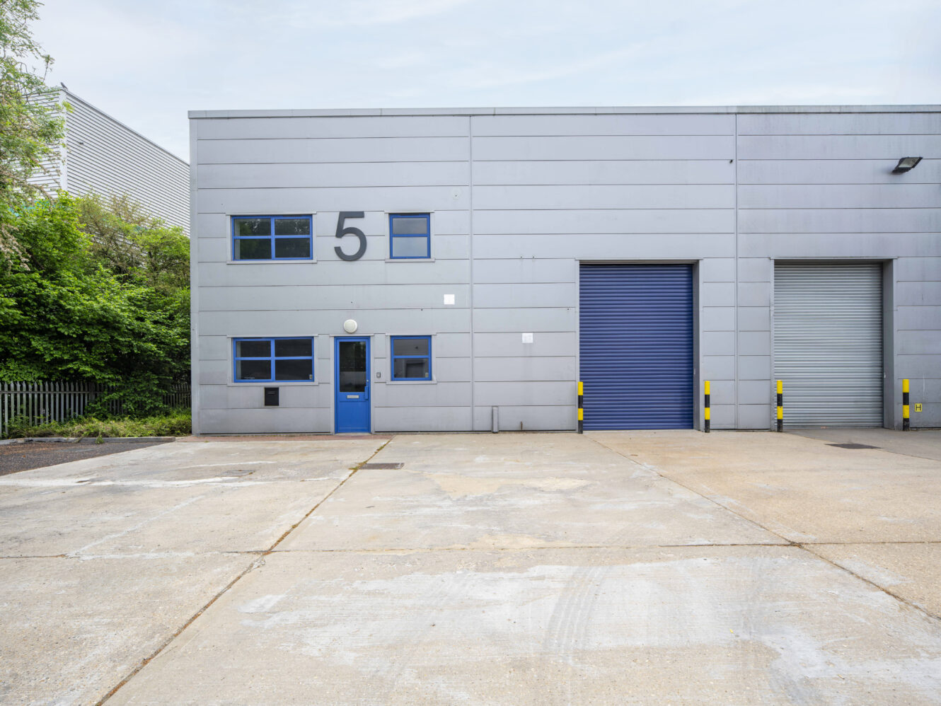 A grey industrial warehouse with a blue door, blue roller shutter, and the number 5 displayed above the door. The area in front is a large, empty concrete lot.