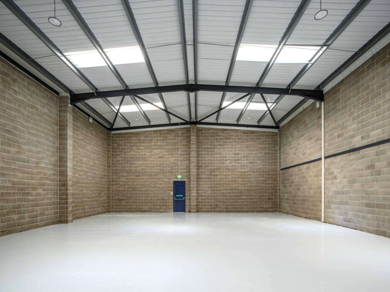 Empty industrial warehouse with exposed brick walls, a gray metal roof, a closed blue door at the back, and a clean white floor.