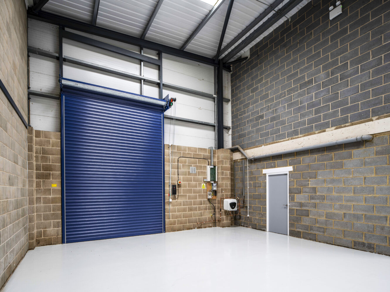 Empty industrial warehouse interior with brick walls, high ceiling, a large blue roller shutter door, grey personnel door, electrical panels, and a clean white floor.