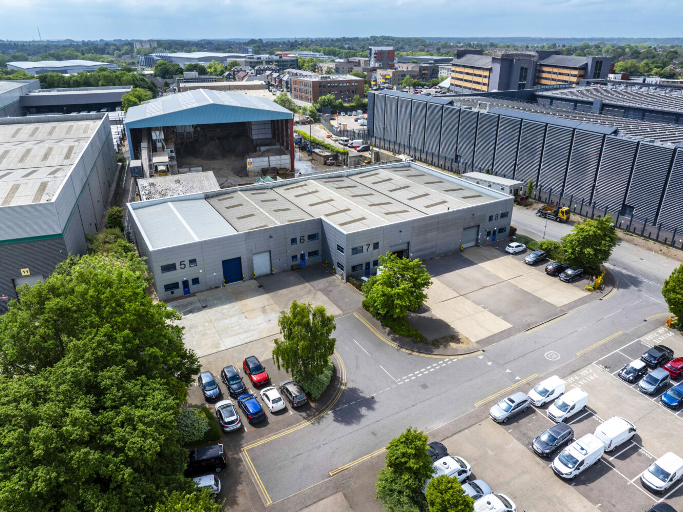Aerial view of an industrial complex with large warehouse buildings, parked cars, and trees, set in an urban area under a clear sky.