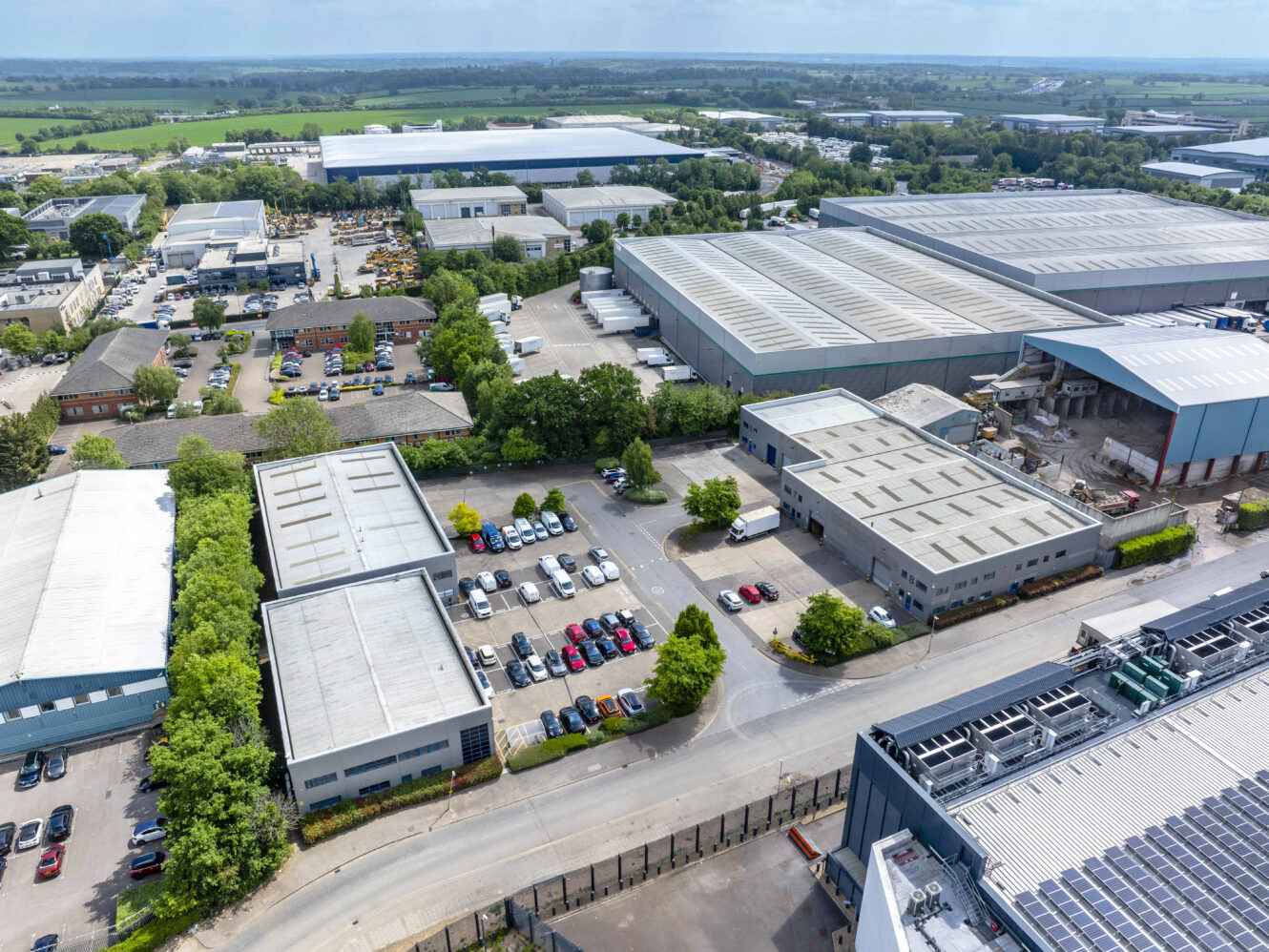 Aerial view of an industrial park with several large warehouses, office buildings, parking lots, and surrounding greenery under a clear sky.