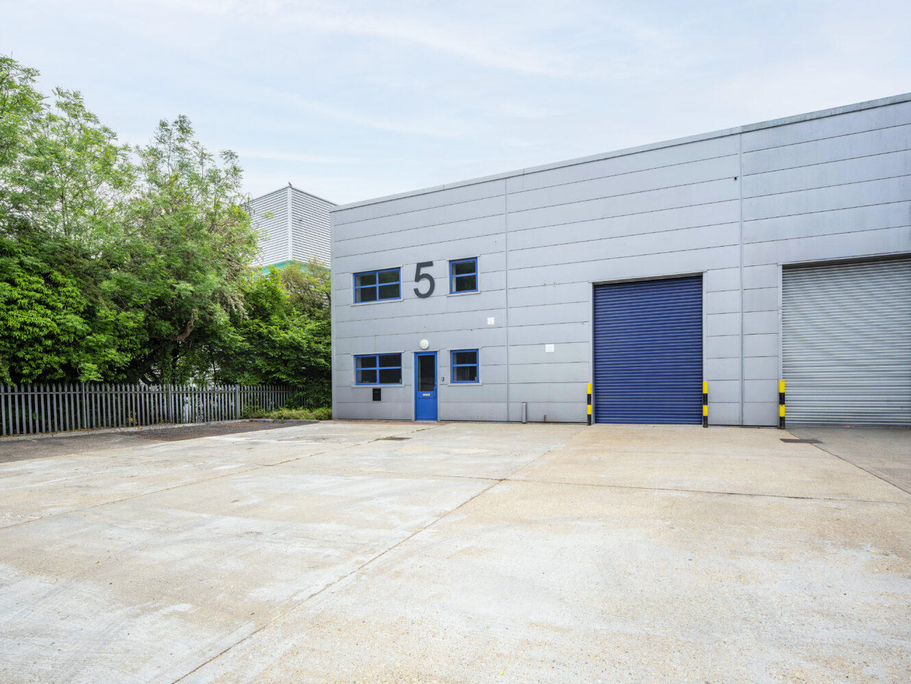 A modern industrial building with a blue door, large blue and grey roller doors, and the number 5 on the facade, set beside a fenced area with green trees.
