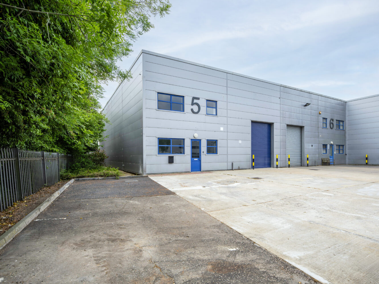 A grey industrial warehouse with blue doors and windows, labeled with a large number 5, sits next to a fenced area and an empty paved lot.