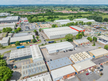Aerial view of an industrial estate with multiple warehouses, parked vehicles, and surrounding green fields under a partly cloudy sky.