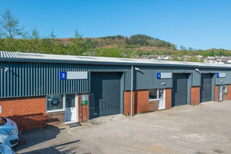 A row of industrial units with blue roller doors and numbered signs, set against a backdrop of trees and hills on a clear day.