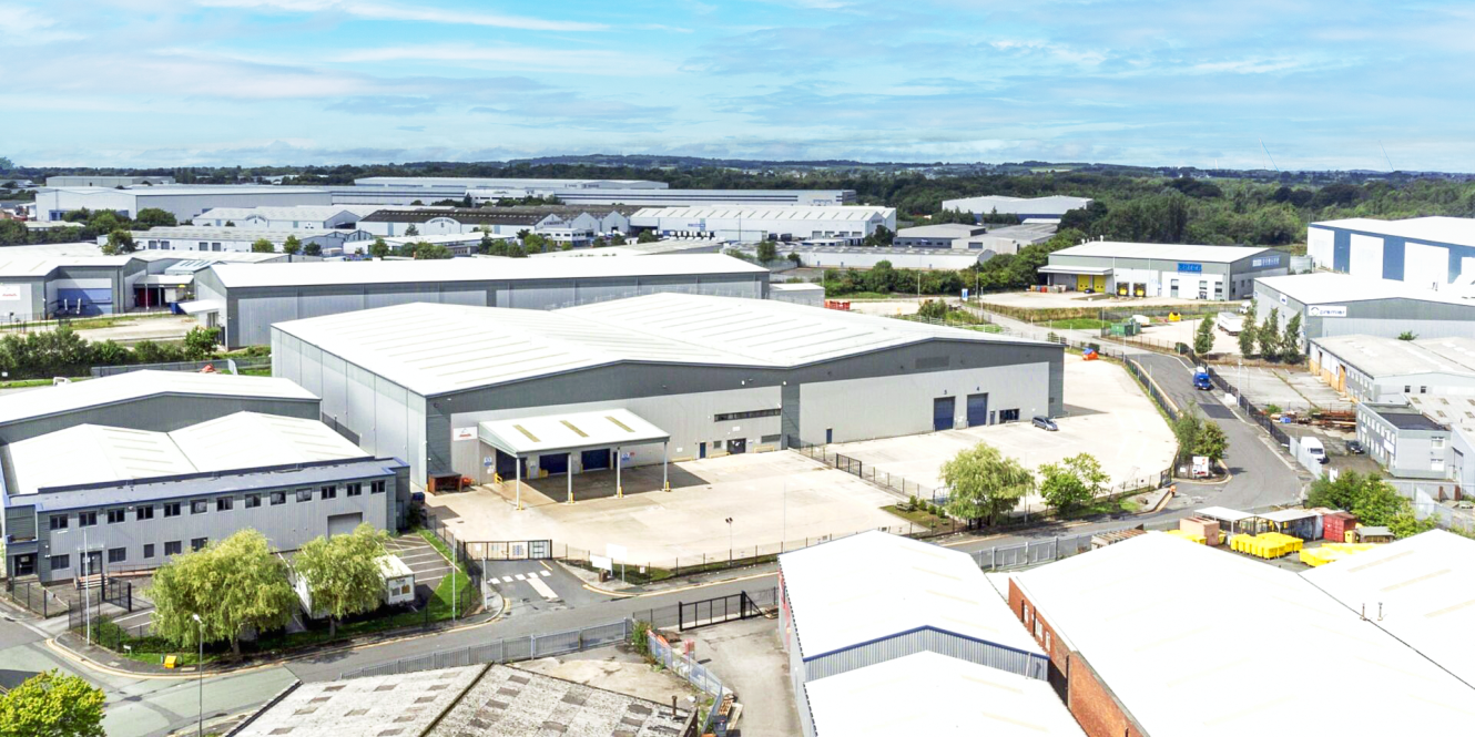 A large industrial warehouse complex with multiple buildings, parking areas, and surrounding roads under a partly cloudy sky.