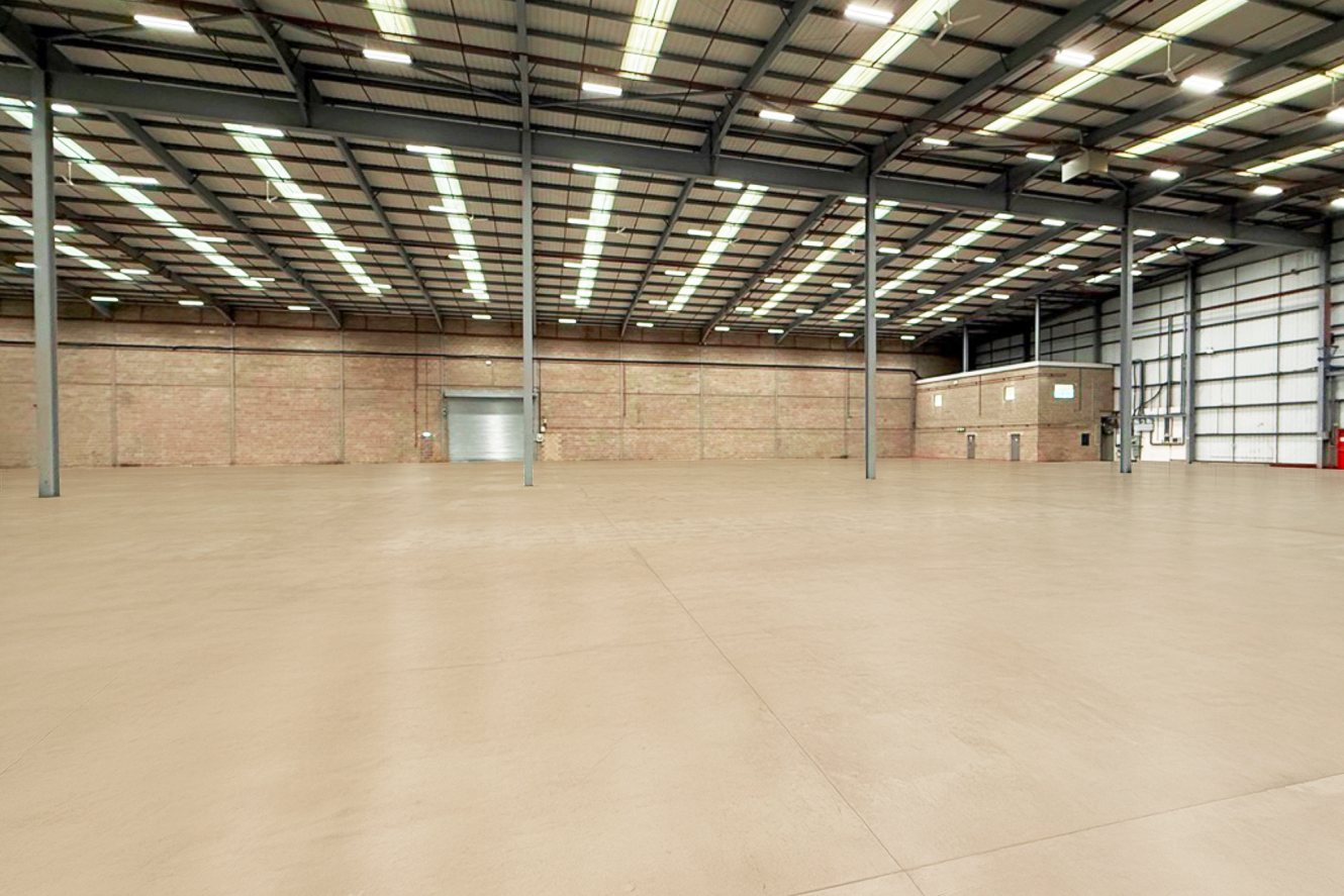 Spacious empty warehouse with high ceilings, exposed brick walls, large overhead lights, and a closed metal roller door.