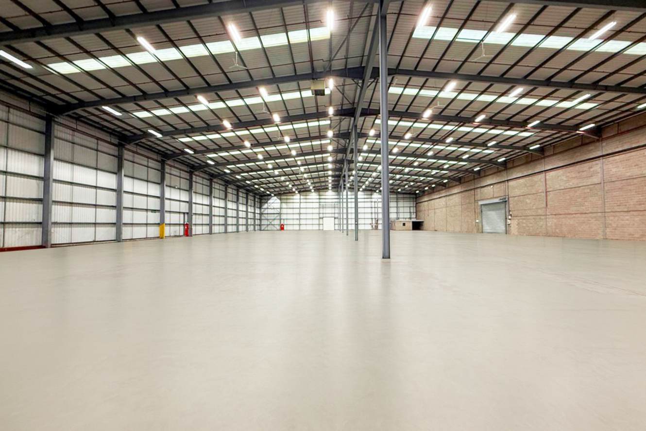 Interior of a large, empty warehouse with high ceilings, metal beams, concrete floor, and fluorescent lighting. Walls are made of corrugated metal and brick.