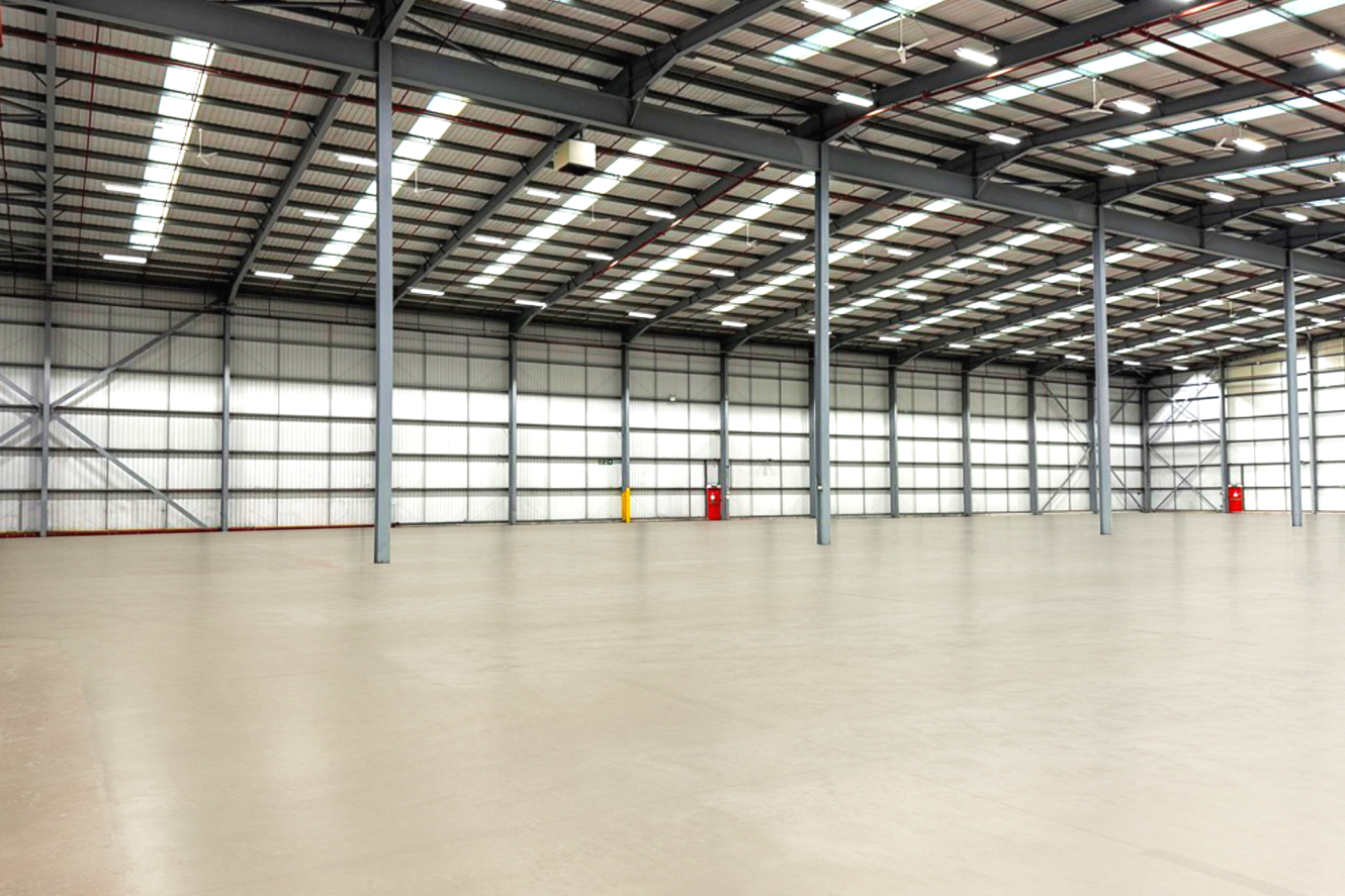 Empty warehouse interior with concrete floor, high metal roof, steel support columns, and large windows letting in natural light.