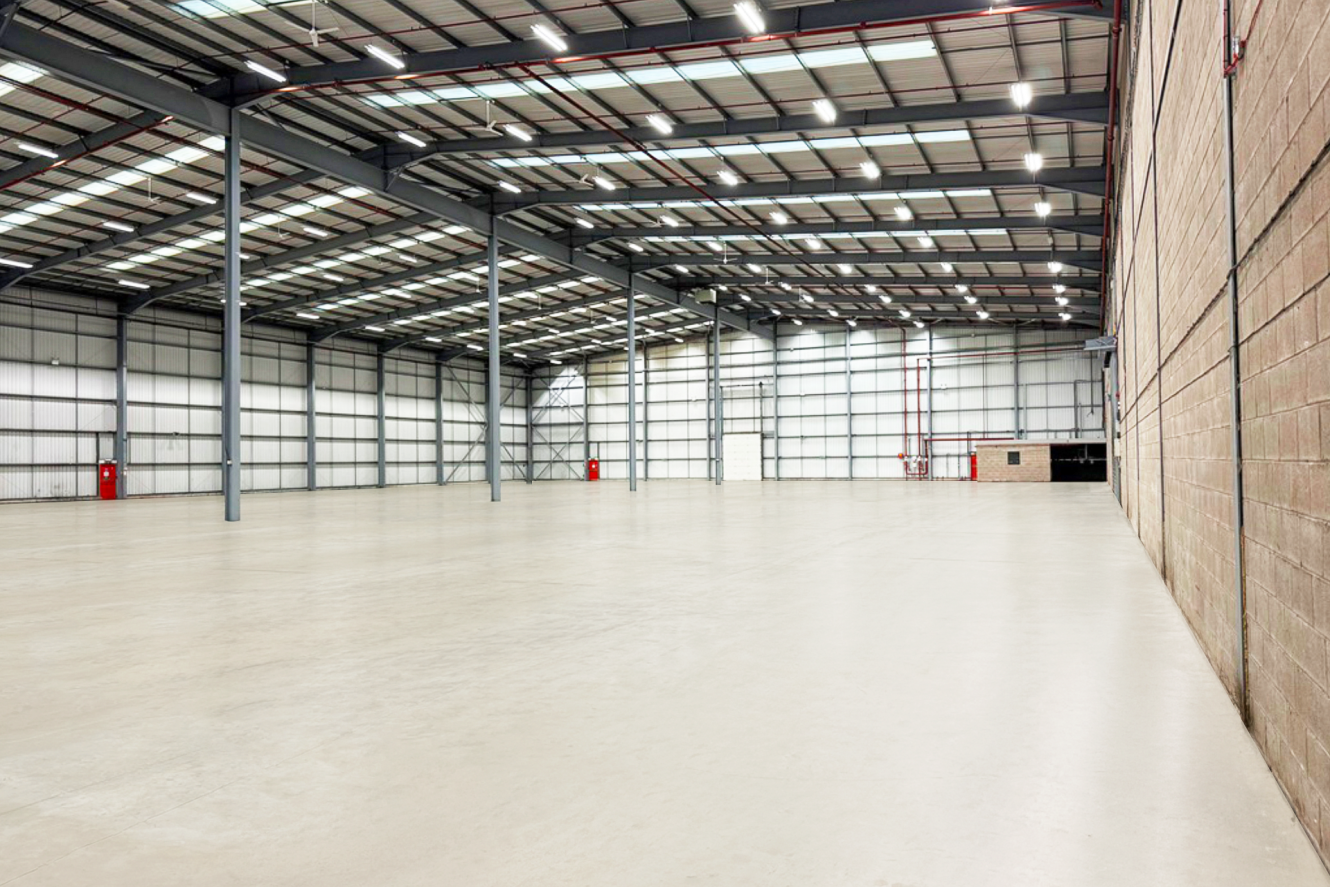 Large empty warehouse with high ceilings, exposed steel beams, concrete floors, and a few red fire extinguishers mounted on the walls.