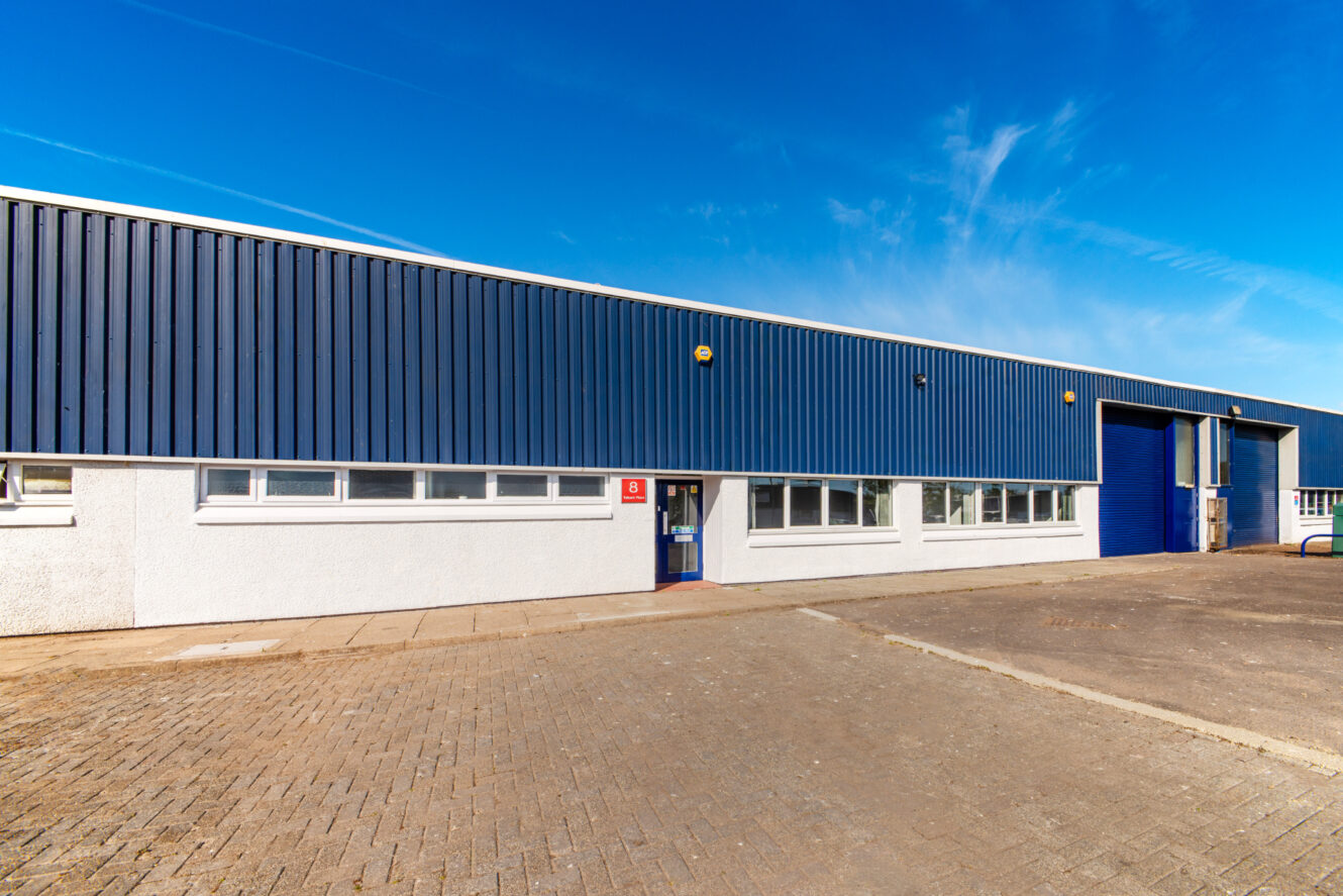 A large industrial warehouse with blue metal siding, white lower walls, small windows, a blue door, and a blue roller shutter under a clear sky.